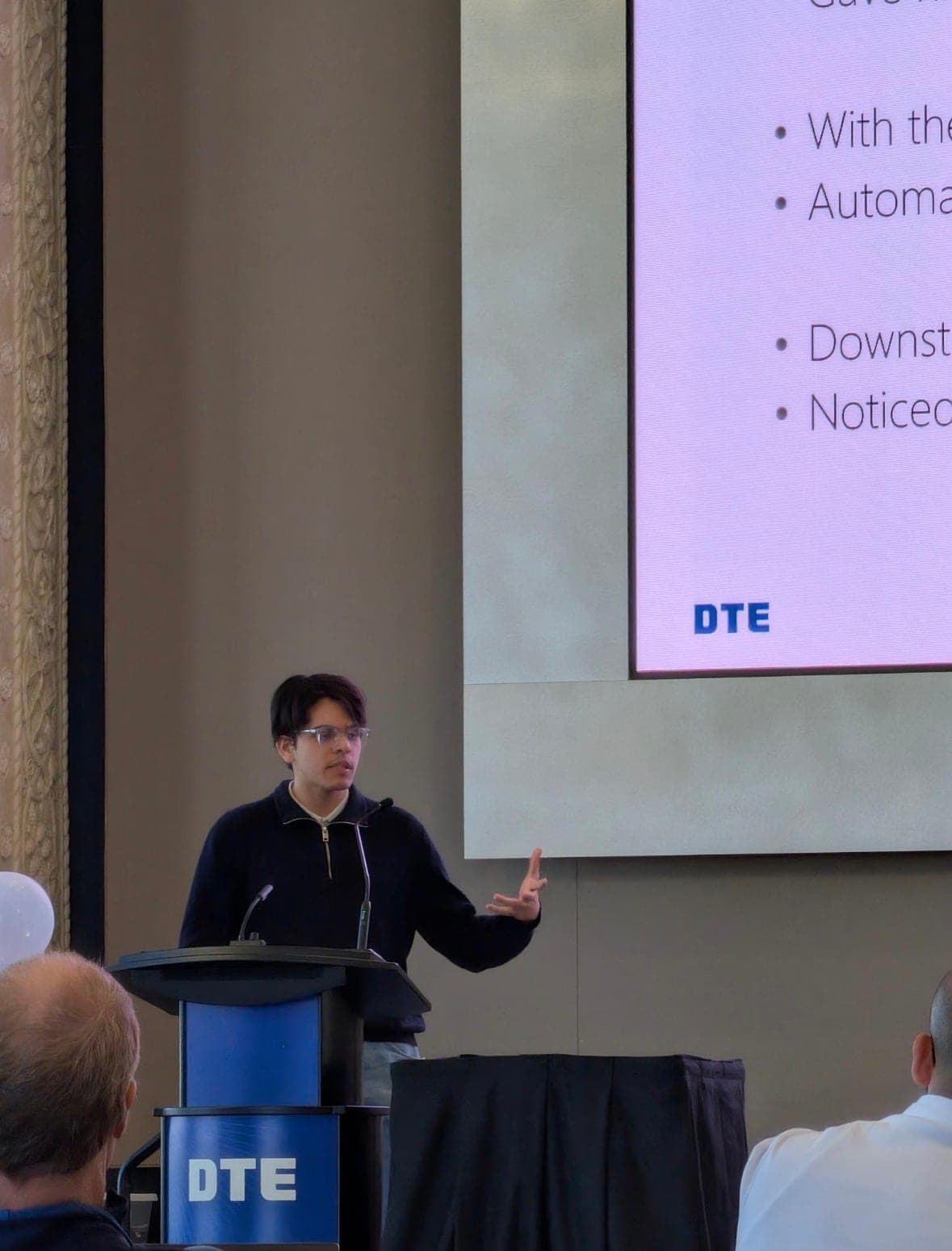 Ahmad presenting at a podium with a DTE-branded slide behind him, listing automation results